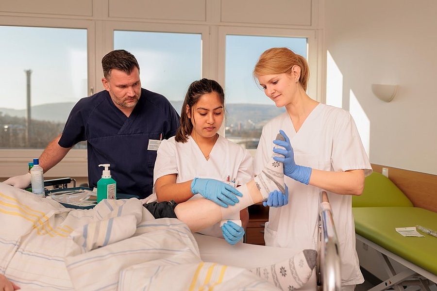 Drei medizinische Fachkräfte in weißer und dunkelblauer Kleidung versorgen einen Patienten im Krankenhausbett, eine trägt blaue Handschuhe und hält den Fuß des Patienten.