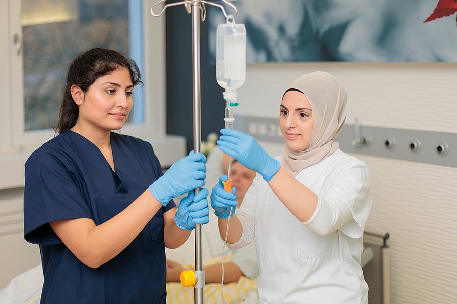 Zwei medizinische Fachkräfte mit Handschuhen stellen gemeinsam einen Infusionsständer mit Tropfbeutel ein.