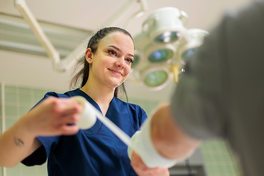Pflegekraft in blauer Arbeitskleidung wickelt einen Verband um einen Arm in einem Behandlungsraum.