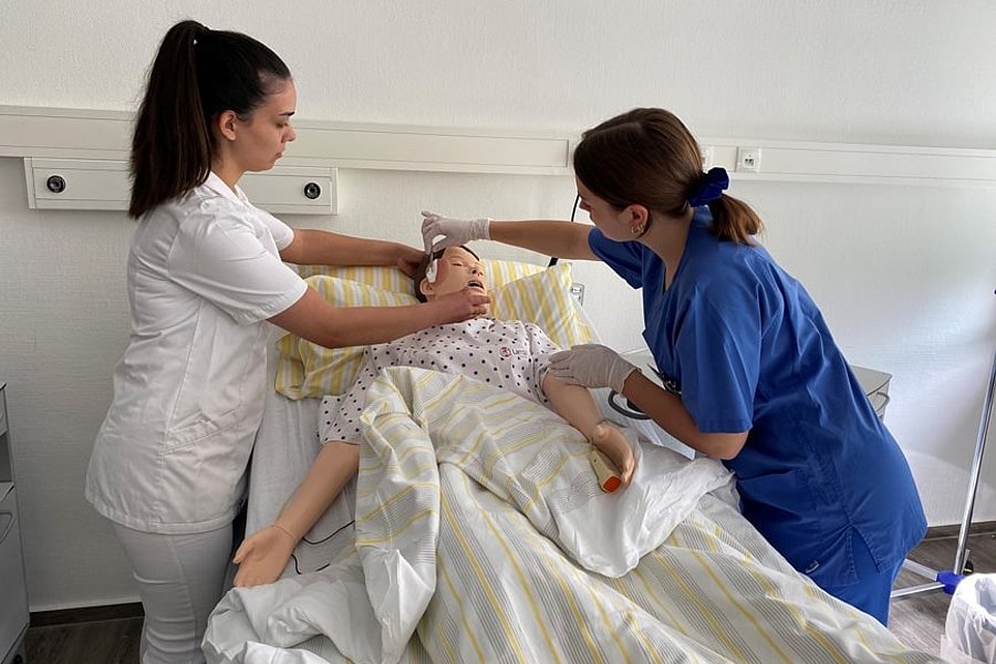 Zwei Pflegekräfte in weißer und blauer Kleidung versorgen eine liegende Person im Krankenhausbett mit gestreifter Bettdecke.