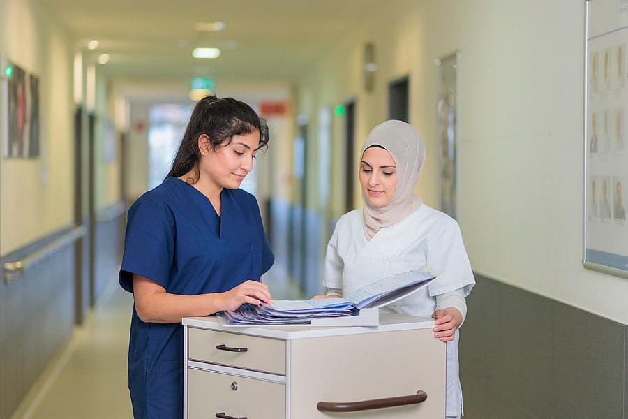 Zwei Pflegekräfte im Flur eines Krankenhauses, eine trägt blaue, die andere weiße Arbeitskleidung und Kopftuch, sie betrachten gemeinsam eine Akte auf einem Rollwagen.