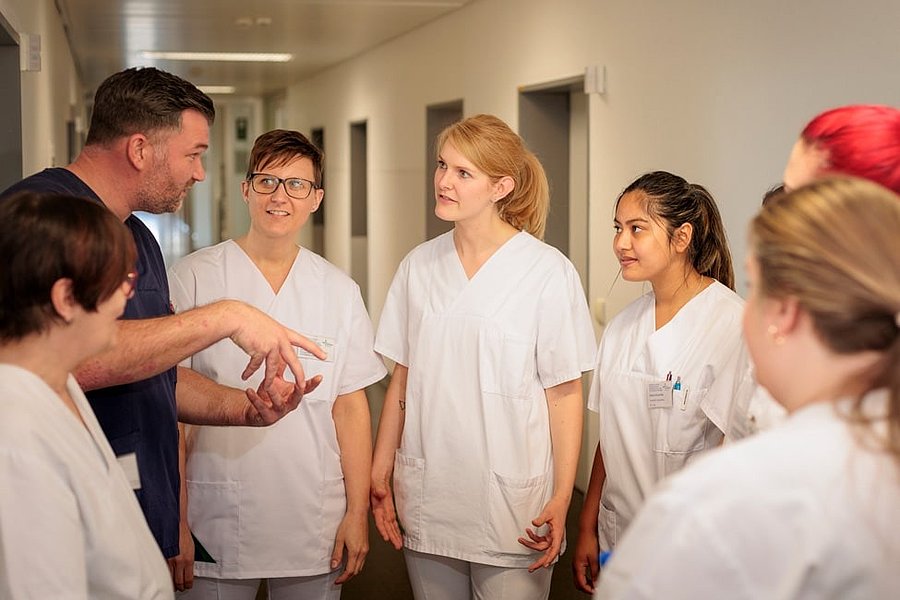 Mehrere Personen in weißen Pflegeuniformen stehen in einem Flur und hören einem Mann zu, der mit den Händen gestikuliert.