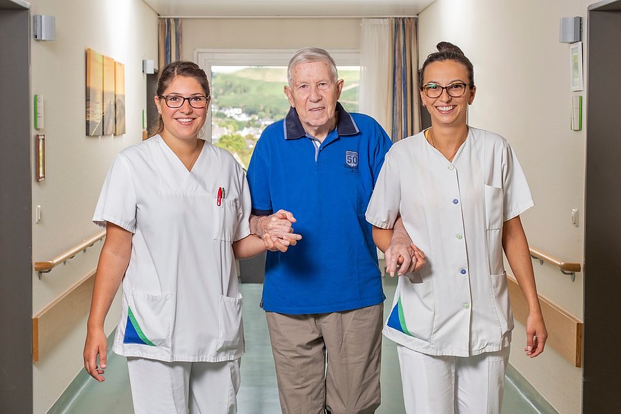 Ein älterer Mann in blauem Poloshirt wird von zwei Pflegekräften in weißer Arbeitskleidung an den Händen gehalten und begleitet.