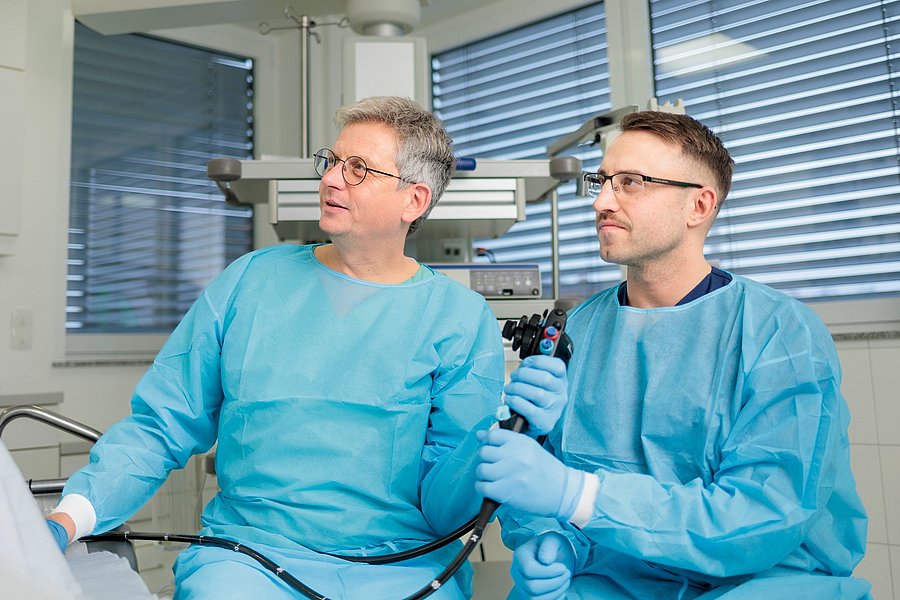 Zwei Personen in blauen Schutzkitteln und Handschuhen halten medizinisches Endoskop in einem Raum mit Fensterjalousien.
