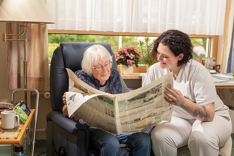Ältere Person sitzt in einem schwarzen Sessel und hält eine Zeitung, neben ihr sitzt eine jüngere Person in weißer Kleidung mit Tätowierung am Unterarm.