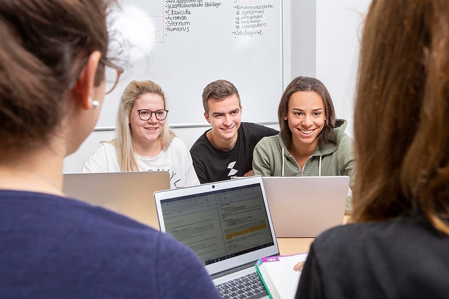 Mehrere Personen sitzen an einem Tisch mit geöffneten Laptops, im Hintergrund eine weiße Tafel mit handgeschriebenen Notizen.