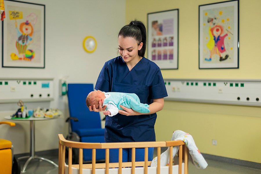 Pflegekraft in blauer Arbeitskleidung hält ein Baby über einem Kinderbett in einem freundlich eingerichteten Raum mit bunten Bildern an der Wand.