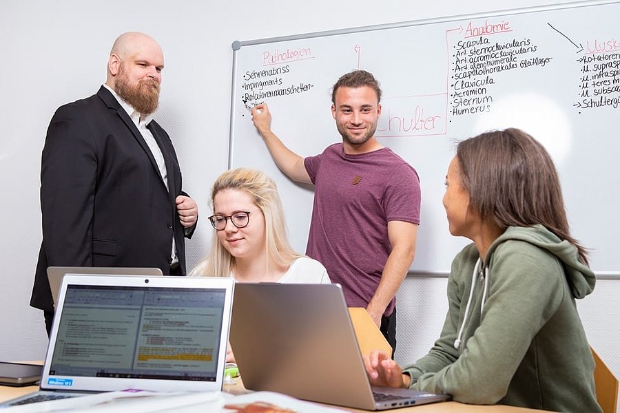 Drei Personen sitzen mit Laptops an einem Tisch, eine Person steht vor einem Whiteboard mit medizinischen Begriffen und zeigt darauf.