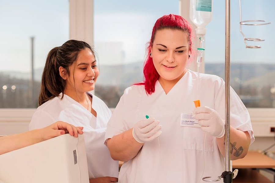 Zwei Pflegekräfte in weißer Arbeitskleidung, eine mit roten Haaren und Handschuhen, neben Infusionsständer im hellen Raum.