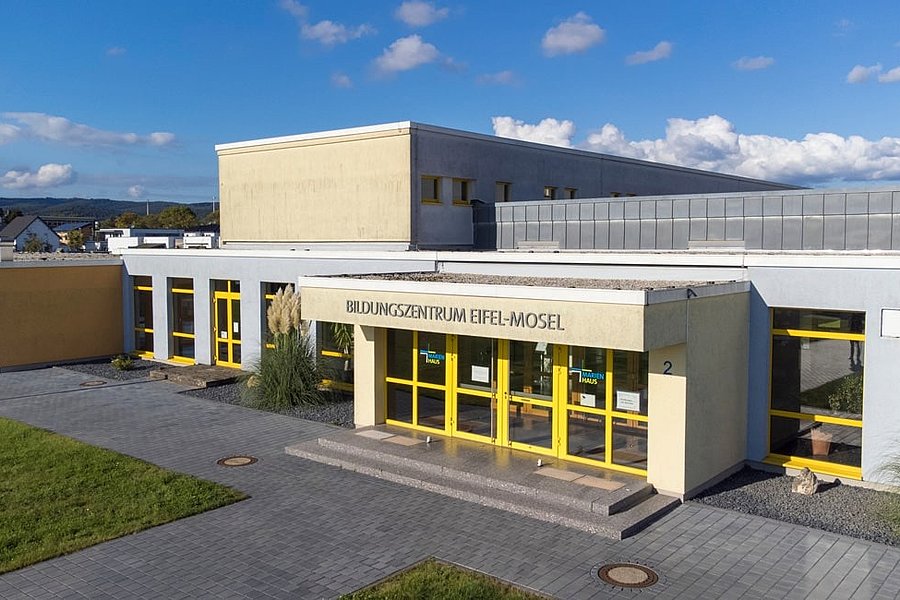 Gebäude mit gelben Türen und Fenstern, Beschriftung 'BILDUNGSZENTRUM EIFEL-MOSEL', davor gepflasterter Weg und Rasenfläche