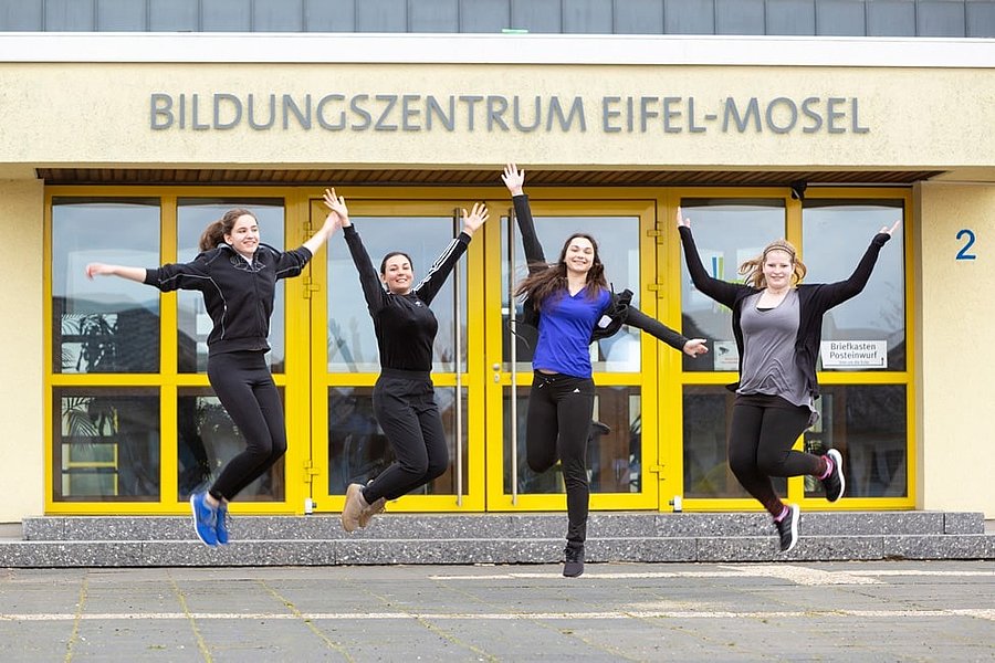 Vier Personen springen vor dem Gebäude mit der Aufschrift 'Bildungszentrum Eifel-Mosel' mit gelben Türen und Fenstern.