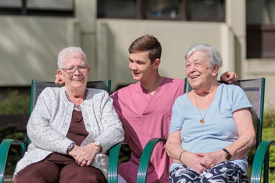 Junge Person in rosa Arbeitskleidung sitzt zwischen zwei älteren Frauen auf Gartenstühlen, alle sitzen nah beieinander im Freien.