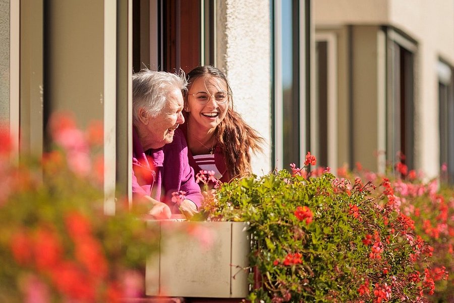 Zwei Personen lehnen an einem Fenster eines mehrstöckigen Gebäudes mit roten Blumen auf dem Balkon.