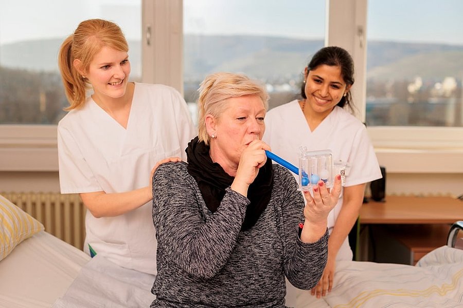 Ältere Frau sitzt auf Bett und hält medizinisches Atemtherapiegerät, zwei Pflegekräfte in weißer Kleidung stehen dahinter.