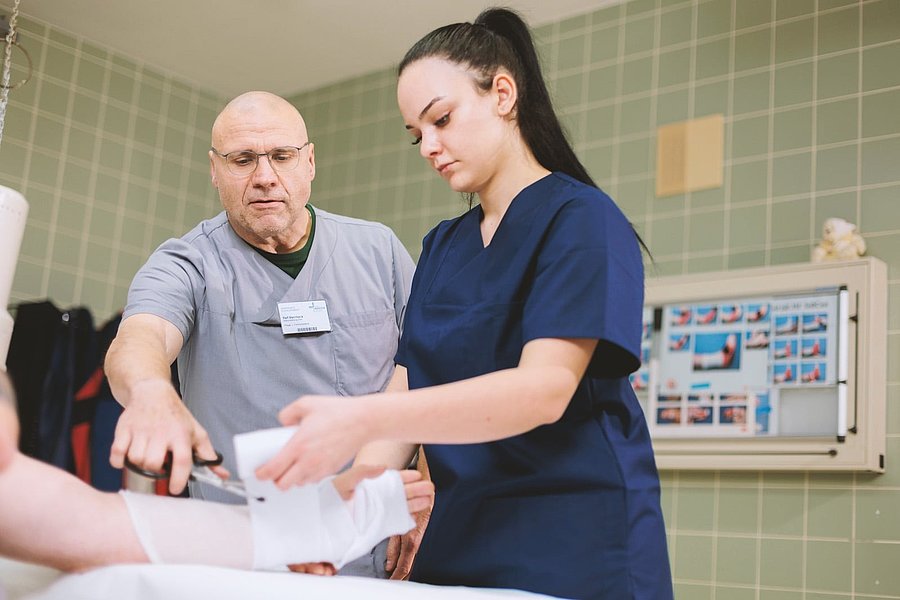 Pflegekraft in blauer Arbeitskleidung wickelt einen Verband um den Unterarm einer Person im Krankenhauszimmer.