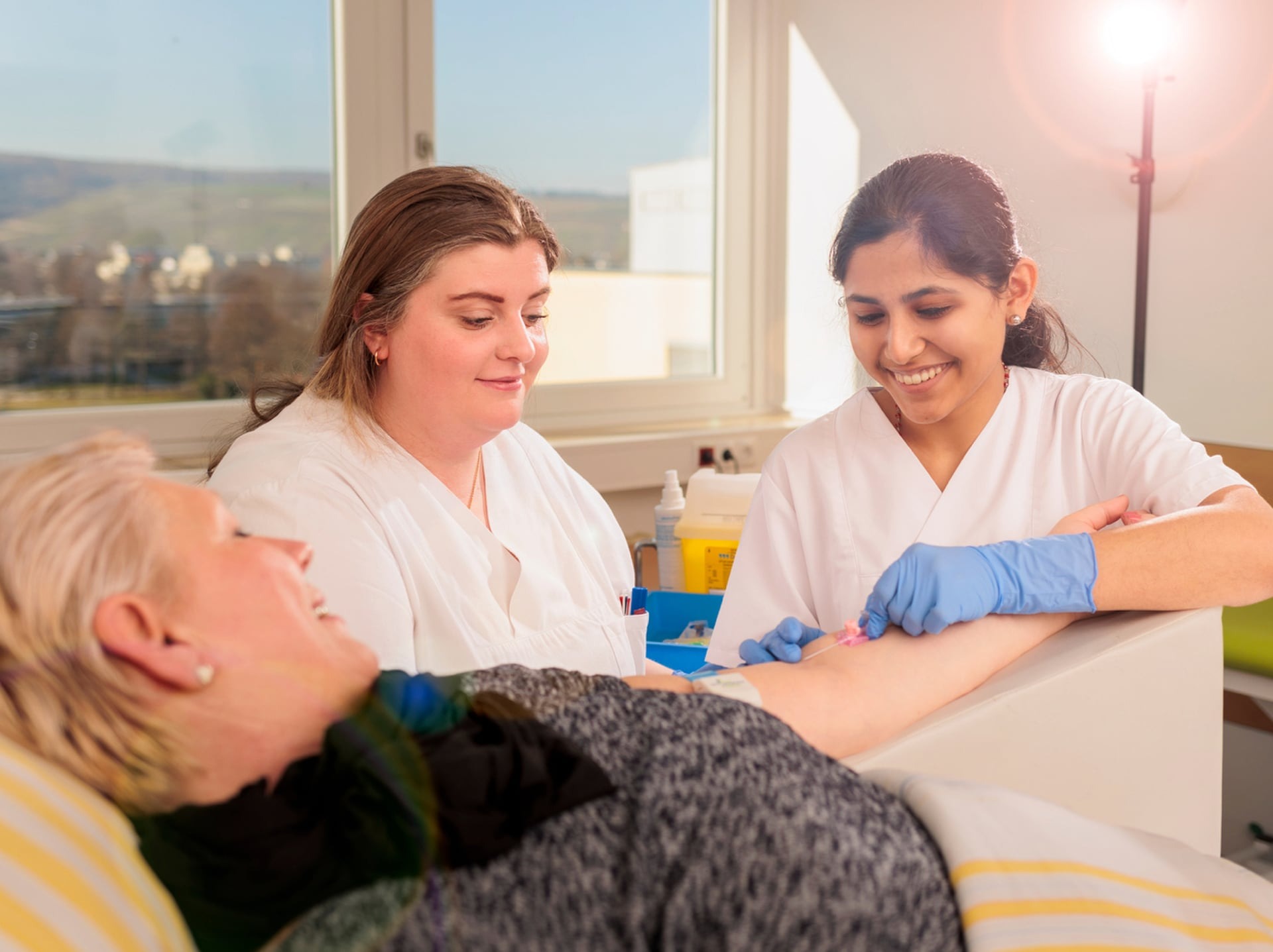 Zwei medizinische Fachkräfte in weißer Kleidung nehmen Blut ab bei einer liegenden Person im hellen Behandlungsraum.